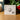 The product packaging box for the 'Mary' Gem V2 by artist Angela Johnson, displayed on a wooden tabletop with a blurred background of green foliage and red berries.