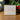 A white square product box for Angela Johnson Sculptures 'Mary V2' sits on a wooden table with a blurred background of green foliage and red berries.