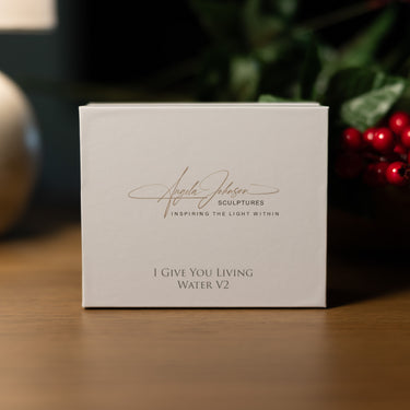 A square white product box standing on a wooden surface, printed with the signature and name Angela Johnson and the text 'I GIVE YOU LIVING WATER V2'. The background is softly blurred with greenery and red berries.
