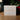 A white square product box standing on a wooden surface, printed with the signature 'Angela Johnson Sculptures' and the text 'I AM THE LIGHT OF THE WORLD V2'. The background is softly blurred with green foliage and red berries.