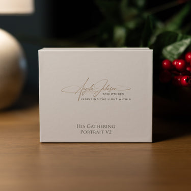 A white square product box standing on a wooden surface, with the front printed with the signature-style text 'Angela Johnson Sculptures' and 'His Gathering Portrait V2'. The background is softly blurred with warm indoor lighting and holiday greenery/red berries.