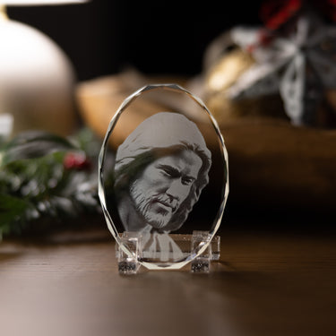 A clear oval crystal plaque featuring an engraved portrait of Jesus Christ, displayed on a small clear stand on a wooden surface with a softly blurred holiday background.