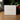 A white square product box with elegant gold and black text reading 'Angela Johnson Sculptures' and 'Because of Love V2', set on a wooden surface with a softly blurred background of greenery and red berries.