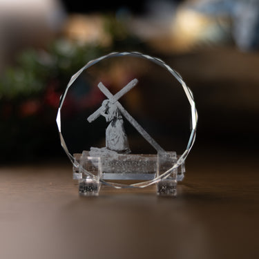 A clear crystal keepsake with an engraved image of Jesus carrying the cross, displayed on a clear acrylic stand on a wooden surface.