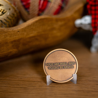 A round bronze-colored metal medallion displayed on a small clear stand on a wooden surface, with engraved text reading 'NOT MY WILL, BUT YOURS BE DONE.' The background is softly blurred and includes a wooden bowl and holiday decorations, crafted by Angela Johnson.