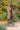 A bronze statue of Jesus Christ standing on a stone base looking up in pray, in Angela Johnson's work O My Father, 3/4 view photographed outdoors among green shrubs and trees.