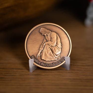 A bronze-toned medallion featuring an embossed kneeling Jesus Christ and the engraved text 'Neither do I condemn you,' displayed on a small clear stand on a wooden surface.