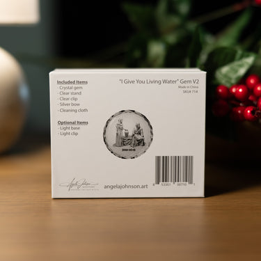 The back of the packaging box for the 'I Give You Living Water' Gem V2 by angelajohnson.art, displayed on a wooden surface with a blurred background of green leaves and red berries.