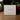 A square white product box standing on a wooden surface, printed with the signature and name Angela Johnson and the text 'I GIVE YOU LIVING WATER V2'. The background is softly blurred with greenery and red berries.