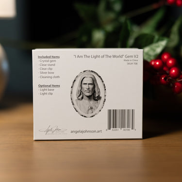 The product packaging box for 'Angel Johnson Art - I Am The Light of The World' Gem V2, displayed on a wooden surface with a blurred background of green foliage and red berries.