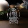 A clear oval crystal plaque featuring a grayscale portrait engraving of Jesus Christ, displayed on a clear acrylic stand on a wooden surface with a softly blurred background.