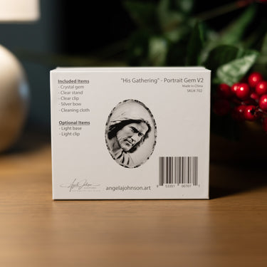 A white product box for a gemstone portrait keepsake, photographed on a wooden surface with blurred greenery and red berries in the background. The box shows the title "His Gathering" - Portrait Gem V2, an oval portrait illustration, included and optional items, and the brand name angelajohnson.