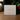 A white square product box standing on a wooden surface, with the front printed with the signature-style text 'Angela Johnson Sculptures' and 'His Gathering Portrait V2'. The background is softly blurred with warm indoor lighting and holiday greenery/red berries.
