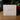 A square off-white product box standing on a wooden surface, printed with the signature 'Angela Johnson SCULPTURES' and 'FIRST VISION V2'. The background is softly blurred with green foliage and red berries.
