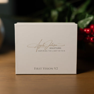 A square off-white product box standing on a wooden surface, printed with the signature 'Angela Johnson SCULPTURES' and 'FIRST VISION V2'. The background is softly blurred with green foliage and red berries.