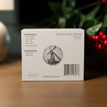 The back of a white product box for a crystal gem, featuring the text 'Because of Love' Gem V2' and a circular illustration of a windmill. The box is placed on a wooden surface with a blurred background of green foliage and red berries.