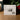 The back of a white product box for 'As One Who Serves' Gem V2, displayed on a wooden surface with a blurred background of greenery and red berries. The box lists included items such as a crystal gem, clear stand, clear clip, silver box, and cleaning cloth, and features a small black-and-white product photo.
