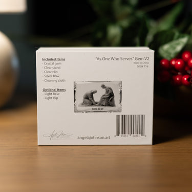 The back of a white product box for 'As One Who Serves' Gem V2, displayed on a wooden surface with a blurred background of greenery and red berries. The box lists included items such as a crystal gem, clear stand, clear clip, silver box, and cleaning cloth, and features a small black-and-white product photo.