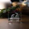 A clear rectangular crystal plaque on a small clear stand, featuring a white relief sculpture of Jesus cleaning his apostles feet and the inscription 'LUKE 22:27' on the front, displayed on a wooden surface with a softly blurred indoor background.