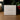 A white square product box standing on a wooden surface, printed with the script logo 'Angela Johnson Sculptures' and the text 'Lazarus, Come forth Jesus, Mary V2'. The background is softly blurred with warm indoor lighting and holiday greenery with red berries.