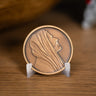 A round bronze-toned medallion with an embossed profile portrait of Jesus Christ, displayed on a small clear stand on a wooden surface.