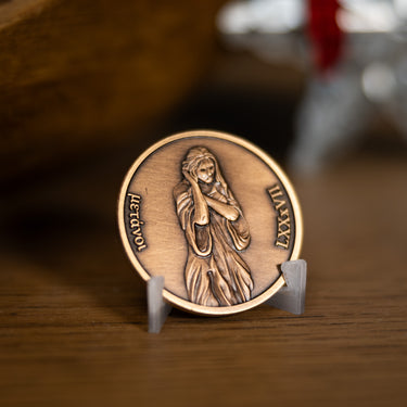 A bronze-toned religious medallion featuring an embossed figure in prayer, based on Angela Johnson's work Neither Do I Condemn, displayed on a small clear stand on a wooden surface with a softly blurred background.