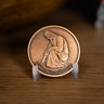 A bronze-toned medallion featuring an embossed kneeling Jesus Christ and the engraved text 'Neither do I condemn you,' displayed on a small clear stand on a wooden surface.