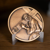 A circular bronze-toned religious medallion featuring a raised relief of Jesus carrying a cross, based on Angela Johnson's wokr Because of Love, displayed on a small clear stand on a wooden surface. 