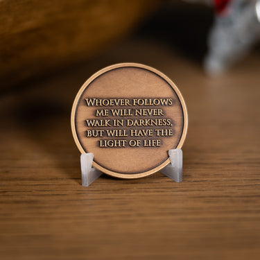 A round bronze-colored metal plaque with an engraved quote, displayed on a small clear plastic stand on a wooden surface, based on Angela Johnson's work Come Unto Me.