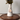 A white ceramic vase with a tapered shape, holding dried green foliage, sits on a dark wood tray against a textured light gray wall. A round pendant with a copper-toned medallion and a rope chain hangs from the vase part of Angela Johnson's Niether Do I Condmen You work.
