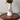 A white ceramic vase with a narrow neck, holding dried green leaves, sits on a dark wood octagonal tray against a light textured wall. A round bronze-toned medallion with a depiction of Jesus Christ hangs from a rope around the vase, based on Angela Johnson's work Come Unto Me.