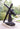 A dark bronze sculpture of Jesus Christ carrying a large wooden cross, mounted on a stone base in Angela Johnson's work Because of Love, in an outdoor garden setting with trees and a pathway visible.