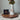 A white ceramic vase with a narrow neck, holding dried green leaves, sits on a dark wood octagonal tray against a light textured wall. A round bronze-toned medallion with a depiction of Jesus Christ hangs from a rope around the vase, based on Angela Johnson's work Come Unto Me.