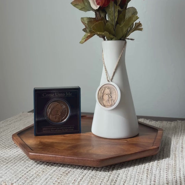 A white ceramic vase with a narrow neck, holding dried green leaves, sits on a dark wood octagonal tray against a light textured wall. A round bronze-toned medallion with a depiction of Jesus Christ hangs from a rope around the vase, based on Angela Johnson's work Come Unto Me.