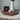A white ceramic vase with a narrow neck, holding dried green leaves, sits on a dark wood octagonal tray against a light textured wall. A round bronze-toned medallion with a depiction of Jesus Christ on clear stand in front of the vase, based on Angela Johnson's work Come Unto Me.