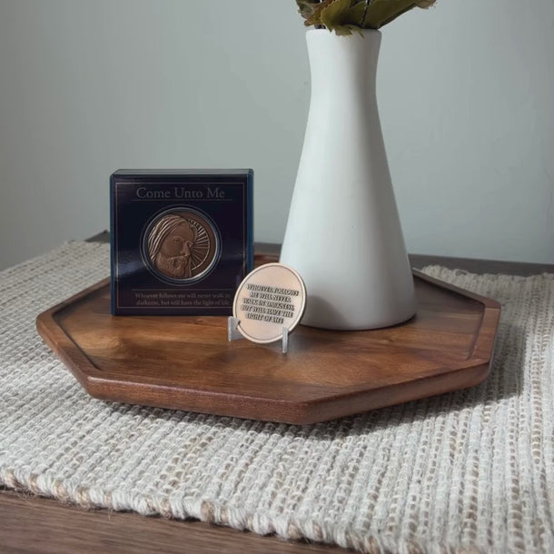 A white ceramic vase with a narrow neck, holding dried green leaves, sits on a dark wood octagonal tray against a light textured wall. A round bronze-toned medallion with a depiction of Jesus Christ on clear stand in front of the vase, based on Angela Johnson's work Come Unto Me.