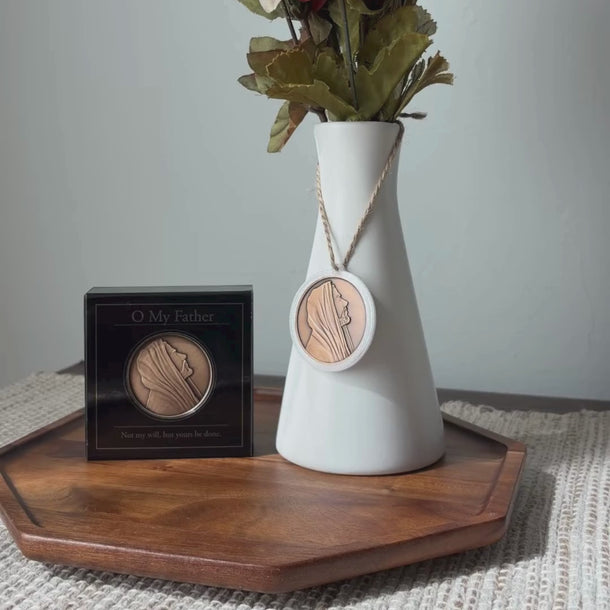 A white ceramic vase with dried green leaves in the neck, hanging from a rope pendant with a round bronze medallion featuring a profile portrait of Jesus Christ from Angela Johnson titled O My Father.